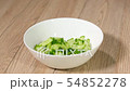 The cook puts sliced cucumbers in a plate and pours gazpacho. 54852278