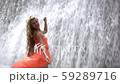 Young happy woman in orange dress standing under spray of water in big waterfall. Wet smiling girl. 59289716