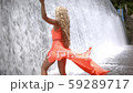 Young woman in dress standing under spray of water in big waterfall. Wet girl rising hands up 59289717