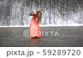 Young woman posing near big waterfall. Girl in orange dress rising hands up. View from back. 59289720