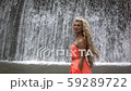 Young woman in orange dress walking and seriously looking in camera on background of big waterfall. 59289722