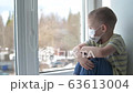 Sad child in protective medical masks sits on windowsill and looks out window. Quarantine, staying at home. Virus protection, pandemic, coronavirus, covid-19, prevention epidemic. 63613004