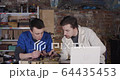 Two young student scientists working in an old dirty workshop garage with some electronics soldering and watching instructions on laptop while speaking to each other 4K 64435453