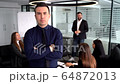Confident young man holding arms folded while standing at the table with talking colleagues on a background 64872013
