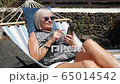 Senior woman looking on cell phone relaxing on a hammock in home garden, fuerteventura spain 65014542