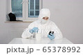Portrait of young male person stitting behind white dining desk in kitchen in protective suit with a cup of tea and reading news on his phone with a black cat in background during pandemic coronavirus 65378992