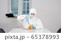 Portrait of young male person stitting behind white dining desk in kitchen in protective suit cutting an orange to eat later with a black cat in background during pandemic coronavirus quarantine time 65378993