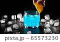 Blue cocktail. Hand puts an orange slice on the edge of glass with blue summer cocktail on the black table with ice cubes on it against black background. Concept of alcohol cocktail party. 4K 65573230