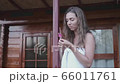 A beautiful girl puts pink flower behind her ear. She is dressed in a white towel and stands outside close to home. The girl looks into the frame and smiles. Medium shot 66011761