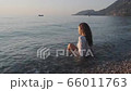 Beautiful young woman in a white blouse sitting on the shore. Happy woman tourist smiling enjoys the warm waves at sunset. Beautiful young woman relaxing on coast during summer vacation. Legs of girl  66011763