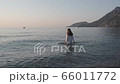 Vacation travel bikini woman walking on beach into water. Beautiful young woman in a white wet blouse in sea water. Happy woman tourist smiling enjoys the warm waves at sunset 66011772