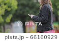 Girl in black jacket walks down the street looking at her mobile phone. 66160929