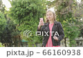 Funny young attractive girl using smartphone and smiles on background of green trees in summer. Pretty girl in a pink sweater writes a message on the phone in the middle of the park. 66160938