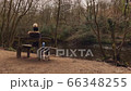 A young woman sitting beside a river in the woods.  An attractive young lady sitting on a wooden bench in the middle of the woods overlooking a river 66348255