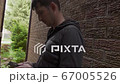 young man who is texting messages on his cellphone standing in a shade of the red bricks wall 67005526