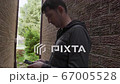 young man who is texting messages on his cellphone standing in a shade of the red bricks wall 67005528
