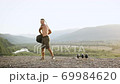 Front view of handsome muscular shirtless tall young man in brown shorts which doing power exercises with tire from wheel on the mount with beautiful nature around 69984620