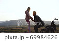 Healthcare family concept where tall incapacitated young man with unusual hairdo trying to get up from wheelchair with his wife's help and then they hugging on the hill with surrounding beautiful 69984626