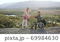 Side view of good-looking smiling careful blondie which helping to stand up from wheelchair her temporarily incapacitated husband and they hugging on the hill surrounded with wonderful nature 69984630