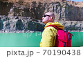 Traveler examines the area. View from the back. Stylish guy with a red backpack dressed in a yellow raincoat stands against the background of a blue lake in a stone canyon. 70190438