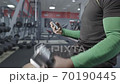Unidentified black man looks at the phone in the gym while lifting a dumbbell. Sport facilities on the background. Smartphone on hands. 70190445