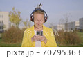 Stylish adult woman in headphones with phone walks along city street on sunny day. 70795363