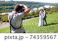 Professional wedding photographer taking pictures of the bride and groom on a hillside covered with green grass 74359567