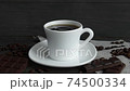 A drop of coffee falls into a white mug of espresso with coffee beans scattered on the table and chocolate on a wooden table. Fresh delicious coffee is ready to drink. 74500334
