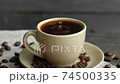 A drop of coffee drops a beige cup of espresso with coffee beans scattered on the table and a linen cloth on a wooden table. Fresh delicious coffee is ready to drink. 74500335