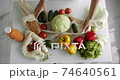 Woman put a cucumbers inside the reusable grocery bag with vegetables on a table with peppers, tomatoes and cucumbers at the kitchen. Zero waste, plastic free, grocery shopping. Mesh cotton shopper. 74640561