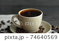 A drop of coffee drops a beige cup of espresso with coffee beans scattered on the table and a linen cloth on a wooden table. Fresh delicious coffee is ready to drink. 74640569