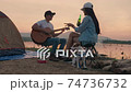 Handheld Young adult Asian couple playing guitar and drinking beer beside their tent campsite while sunset with dog. 74736732
