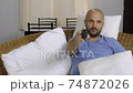 A bearded bald man sits on the couch and switches channels on the TV. 74872026