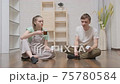 young couple woman and man resting and drinking tea after finishing assembling furniture while sitting on floor in room at home, door of assembled cabinet suddenly falls, they laugh. Home improvement. 75780584