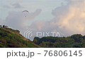 The paraglider soars by parachute on the green mountains. In the distance there is an observation deck with people watching the sunset. 76806145