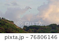 The paraglider soars by parachute on the green mountains. In the distance there is an observation deck with people watching the sunset. 76806146
