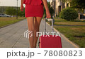 Back view of tourist woman legs in red dress, high heels walking with suitcase in outdoors street city 78080823
