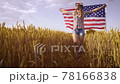 Beautiful young girl holding an American flag on the wind in a field of wheat. Summer landscape against the blue sky. 78166838