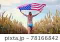 Beautiful young girl holding an American flag on the wind in a field of wheat. Summer landscape against the blue sky. 78166842