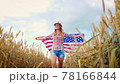 Beautiful young girl holding an American flag on the wind in a field of wheat. Summer landscape against the blue sky. 78166844