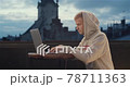 A young man sitting on the roof with a laptop with an old city tower view in the background 78711363