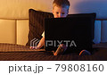 Cute blond Caucasian boy alone in bedroom late at night sitting on bed, typing on laptop keyboard, surfing or trawling Internet. night light in room, moonlight from window. Dolly slides through video. 79808160