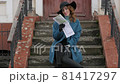 young red-haired woman in a blue coat and black hat with a suitcase on the background of the old city. traveler with map and suitcase 81417297