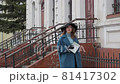 young red-haired woman in a blue coat and black hat with a suitcase on the background of the old city. traveler with map and suitcase 81417302