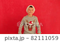 Boy in a New Year's cap and sweater on a red background. Child waiting for Christmas. Happy boy getting ready for the new year. 82111059