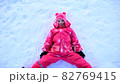 A young girl in a yellow jacket lies in the snow and makes a snow angel with his arms and legs. Child in the snow in winter. Winter outdoor games 82769415