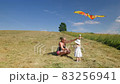 Young grandmother with her grandchildren launches multicolored kite on field with green grass, blue sky and lonely tree 83256941