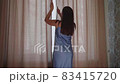 Young woman approaches the window in the bedroom, pushes beige curtains with her hands and enjoys 83415720