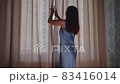 Young woman approaches window, pushes aside curtains and white tulle with hands, opens window and 83416014