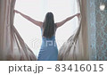 Young woman approaches the window in the bedroom, pushes beige curtains with her hands and enjoys 83416015